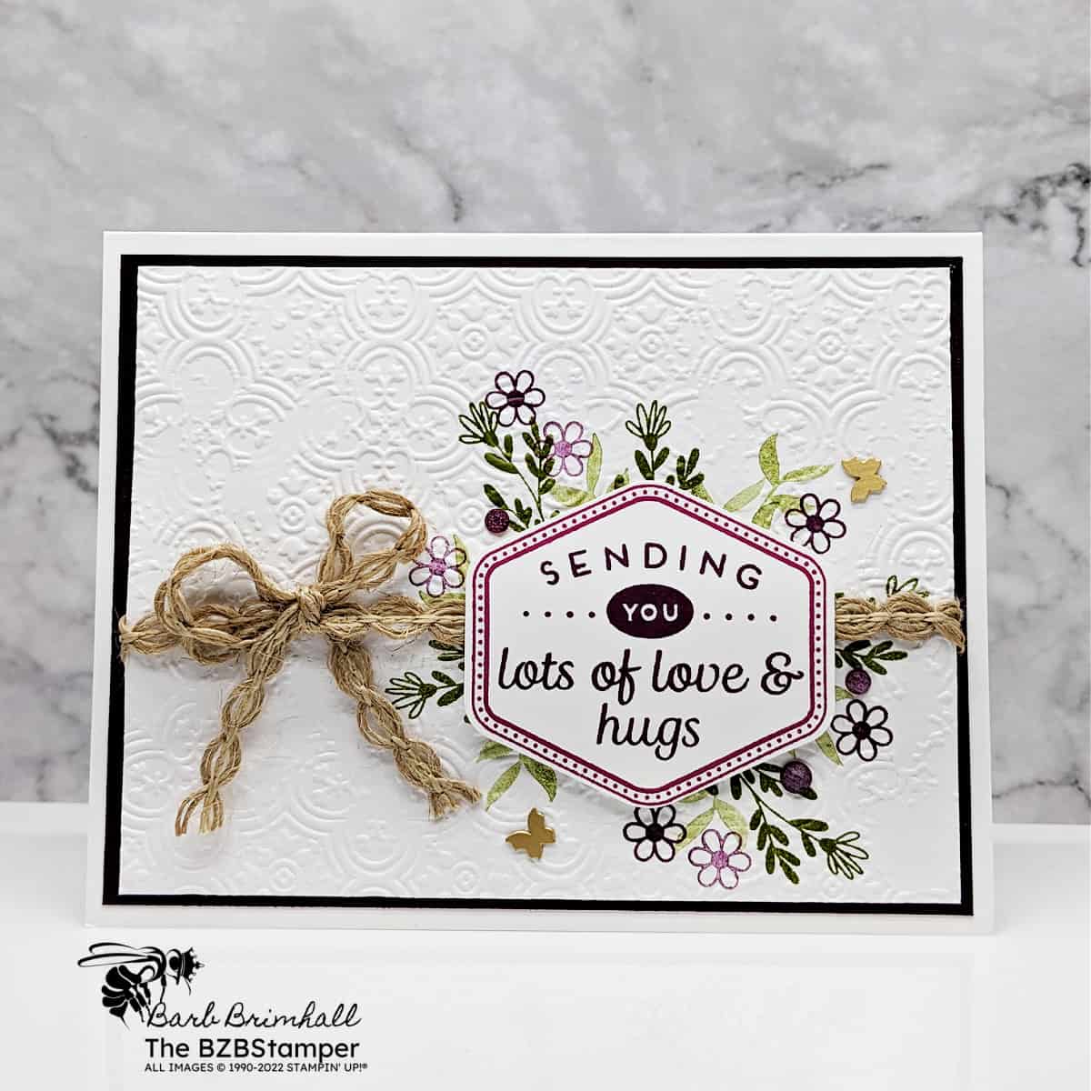Handmade Thinking Of You Card Tutorial featuring purple flowers and green foliage, with an embossed background.  Sentiment is "sending you lots of love & hugs."