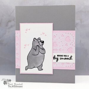 A handmade greeting card with a soft gray background and a light pink accent strip across the center. A white panel features a gray bear standing shyly with small pink hearts floating above its head. The card includes layered pink and white sections, a heart-patterned panel, and a small sentiment strip on the right, creating a sweet and gentle mood.