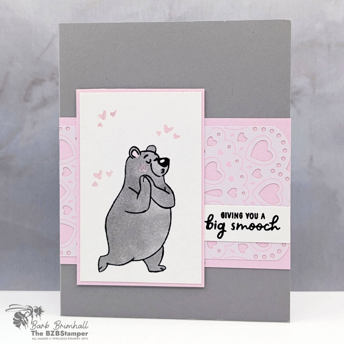 A handmade greeting card with a soft gray background and a light pink accent strip across the center. A white panel features a gray bear standing shyly with small pink hearts floating above its head. The card includes layered pink and white sections, a heart-patterned panel, and a small sentiment strip on the right, creating a sweet and gentle mood.